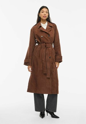 Manteau trench en suede marron avec une taille ceinturée, des boutons double boutonnage et des manches larges. Assorti à un jean gris à jambes larges et des talons aiguilles noirs.