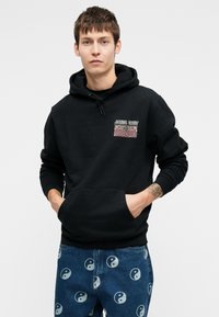 Mustassa hupparissa on etutasku, naruhuppu ja graafinen yksityiskohta. Yhdistettynä sinisiin farkkuihin, joissa on yin-yang-kuvio.