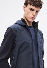 Tumszila hibrīdā jaka ar rāvējslēdzēju priekšpusē, kapuci un teksturētām piedurknēm. Aprīkota ar kontrastējošu audumu, minimālistisku dizainu un smalkām šuvju detaļām.