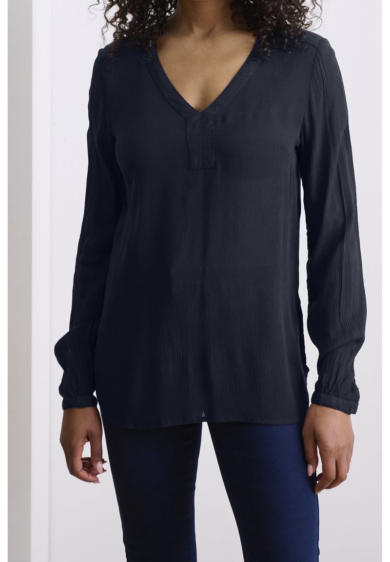 Blouse noire à manches longues avec un col en V, tissu texturé et poignets boutonnés, associée à un pantalon ajusté bleu foncé.