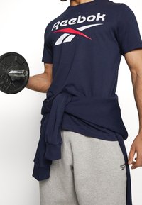 Mörkblå Reebok-t-shirt med vit och röd logo, kombinerad med grå sweatpants. En mörk skjorta knuten runt midjan ger en extra textur.