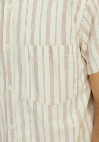 Camisa de manga corta con botones en color crema con delgadas rayas rojas verticales; cuenta con un bolsillo en el pecho izquierdo y una textura de tela suave y ligera.