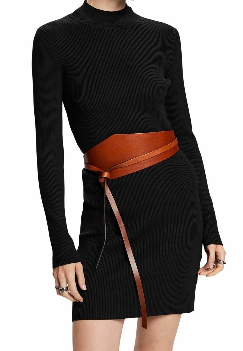 Femme portant une robe noire côtelée ajustée à manches longues avec une large ceinture en cuir marron nouée à la taille, montrant des bagues aux deux mains.