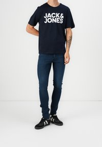 Mężczyzna w czarnej koszulce "JACK & JONES", ciemnoniebieskich skinny jeansach, czarnych sneakersach z białymi paskami, stojący na białym tle.