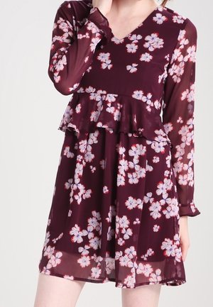 Woman wearing a burgundy floral dress with long sheer sleeves and a ruffled waist detail.
