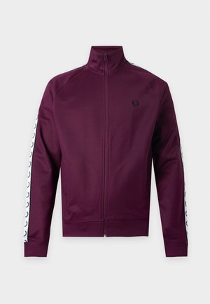 Hombre de pie con chaqueta burdeos con cremallera y rayas blancas con logo en las mangas, pantalones negros, zapatillas blancas y gorra beige.