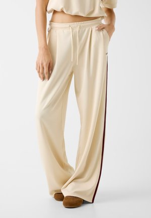 Pantalon beige à jambes larges avec une taille élastique à cordon et des bandes latérales bordeaux, porté avec des chaussures marron et un haut beige court.