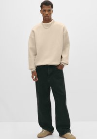 Hanorac oversized bej, cu umeri căzuți, purtat cu blugi negri drepti și adidași maro deschis, având o textură și un design subtil.