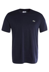 Camiseta de algodón azul marino, de manga corta, con cuello redondo y un pequeño logotipo verde de Lacoste en el lado izquierdo del pecho. Textura suave.