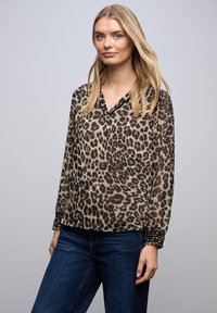 Kvinde med langt blondt hår iført en gennemsigtig leopardprintbluse med lange ærmer og mørkeblå jeans foran en ensfarvet baggrund.