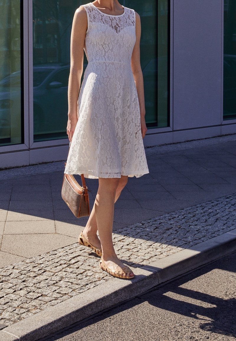Femme debout sur le trottoir portant une robe en dentelle blanche sans manches et des sandales beige, tenant un sac à main marron à ses côtés.