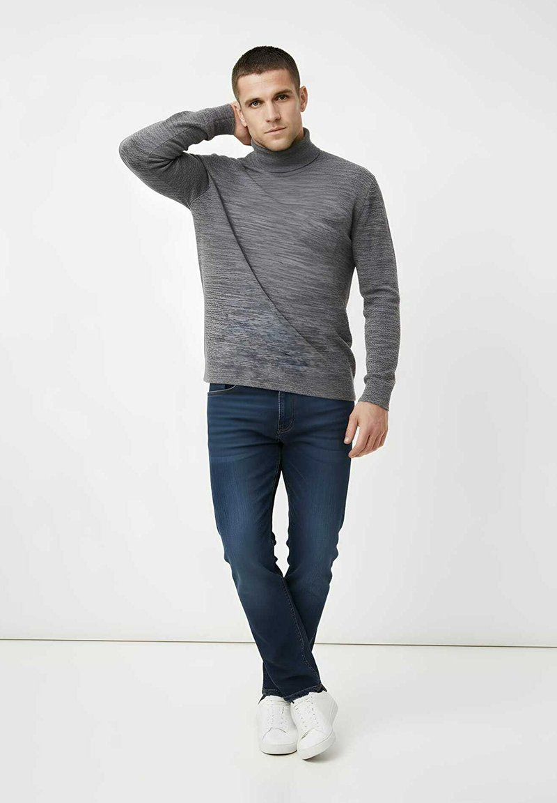 Suéter gris de cuello alto tejido con un patrón texturizado, combinado con jeans ajustados azul oscuro y zapatillas blancas. Posición de pie, fondo neutro.