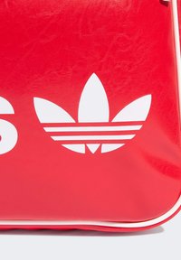Borsa in pelle rossa con logo trefoil bianco e testo in grassetto sul davanti, con una superficie liscia e bordi bianchi a contrasto.