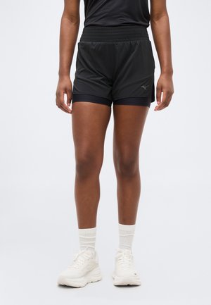 Osoba ubrana w czarne, warstwowe, sportowe spodenki, białe skarpetki i białe tenisówki, stojąca na tle jasnego, jednolitego tła.