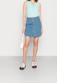 Canotta azzurro chiaro con dettaglio a fiocco, abbinata a una gonna di jeans con chiusura frontale a bottoni e tasche, accessoriata con una borsa bianca.