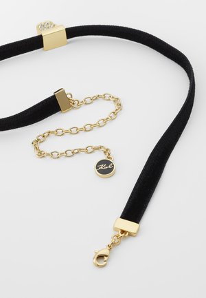 Czarny welurowy naszyjnik na szyję z przedłużką z złotego łańcuszka, zapięciem typu karabińczyk oraz okrągłą zawieszką z wygrawerowanym imieniem "Karl" w stylu pisma odręcznego.