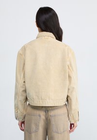 Veste en denim beige avec une coupe décontractée, dotée d'un col pointu et de boutons-pression. L'arrière présente des détails de couture et des nuances de texture usée.