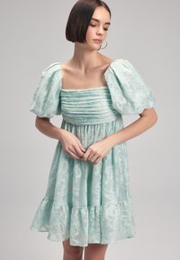 Vaaleansininen mekko, jossa on pullistuvat hihat, rypytetty yläosa ja kukkakuosiset kankaat. Mekossa on myös kerroksellinen röyhelöhelma ja se ulottuu polven ylle.