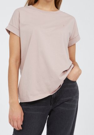Femme portant un t-shirt rose clair à manches courtes avec manches retroussées et un jean noir, une main dans la poche, sur un fond uni.