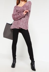 Grober kastanienbrauner Strickpullover mit V-Ausschnitt und asymmetrischem Saum, kombiniert mit schwarzen Skinny-Jeans und schwarzen Stiefeletten. Eine graue Tote-Tasche ist ebenfalls zu sehen.