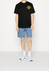 Black cotton T-shirt with yellow Lacoste logo, paired with light blue denim shorts. Model wears white and blue sneakers with white socks.