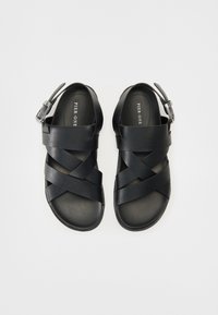 Svarte sandaler med kryssede stropper, laget av teksturert materiale. Har en spenne på ankelstroppen og en flat såle.