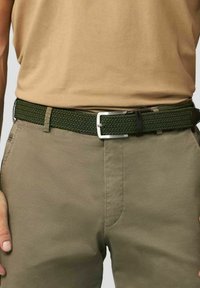 Geweven groene riem met een zilveren rechthoekige gesp. Gepaard met een tan top en khaki broeken, die een getextureerd, gevlochten ontwerp laten zien.