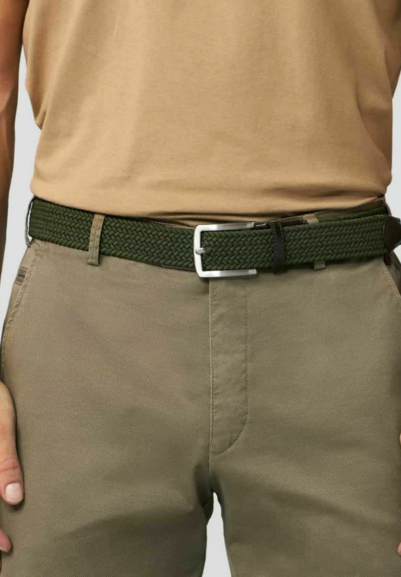 Geweven groene riem met een zilveren rechthoekige gesp. Gepaard met een tan top en khaki broeken, die een getextureerd, gevlochten ontwerp laten zien.
