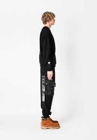 Sudadera negra y pantalones con bolsillo de carga, con detalles gráficos. Botas marrones con suela negra. Perfil lateral recto contra un fondo blanco.