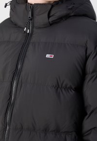 Doudoune noire avec une fermeture éclair frontale et une capuche attachée, arborant un petit logo rectangulaire rouge, blanc et bleu sur la poitrine.