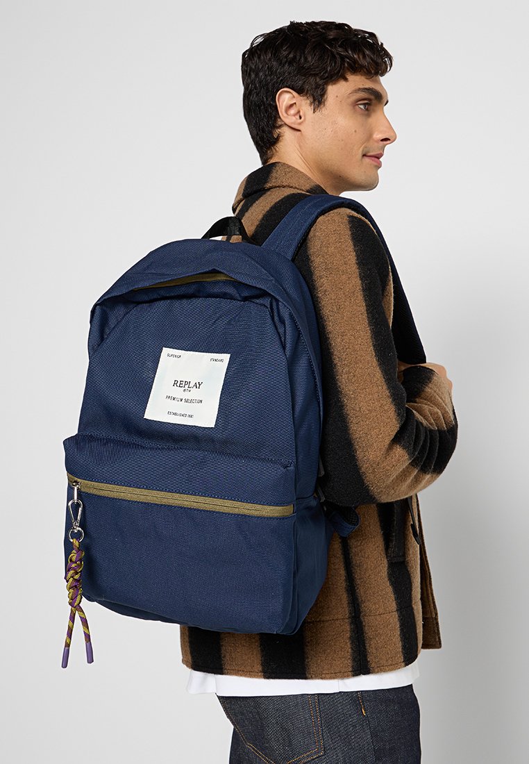 Zaino blu navy con tessuto testurizzato, dettagli con cerniera dorata, tasca frontale e una patch con logo bianco. Ciondolo intrecciato colorato attaccato alla cerniera.