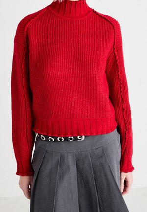 Czerwony sweter z prążkowanym golfem z ozdobnymi szwami na rękawach, w połączeniu z szarą plisowaną spódnicą i czarnym paskiem z dużymi srebrnymi przelotkami.