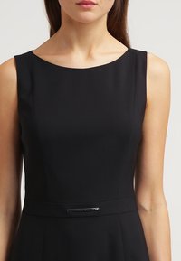 Vestido negro sin mangas con un corte ajustado, con escote redondo y un acento plateado en la cintura. Textura suave y diseño estructurado.