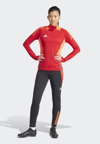 Haut de sport à manches longues rouge et orange avec une demi-fermeture éclair, associé à des leggings noirs avec des accents orange et un logo. Chaussures de foot noires.