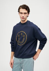 Joven con cabello oscuro que lleva una sudadera azul marino con una cara sonriente amarilla bordada y las letras "H G O", combinada con jeans de color azul claro.