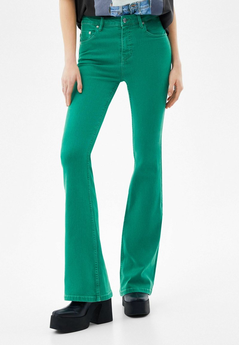 Personne portant un jean bootcut vert taille haute et des chaussures plateforme noires, debout devant un fond blanc.