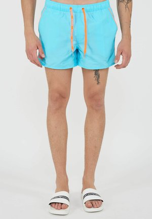 Man draagt lichtblauwe zwembroek met oranje trekkoorden en witte slippers, toont tatoeages op linkerarm en rechterdij.