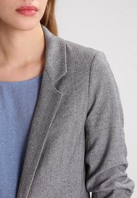 Szary blazer o teksturze z subtelnym wzorem w jodełkę i wykrojnymi klapami, noszony na niebieskiej bluzce z delikatnym połyskiem.