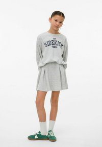 Sweatshirt gris avec le graphisme "MON PARTENAIRE", associé à une jupe plissée grise. Des chaussettes blanches et des baskets vertes complètent la tenue.