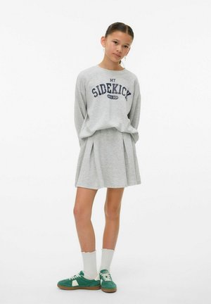 Grå sweatshirt med "MIN SIDEKICK" grafik, parret med en plisseret grå nederdel. Hvide sokker og grønne sneakers fuldender outfittet.