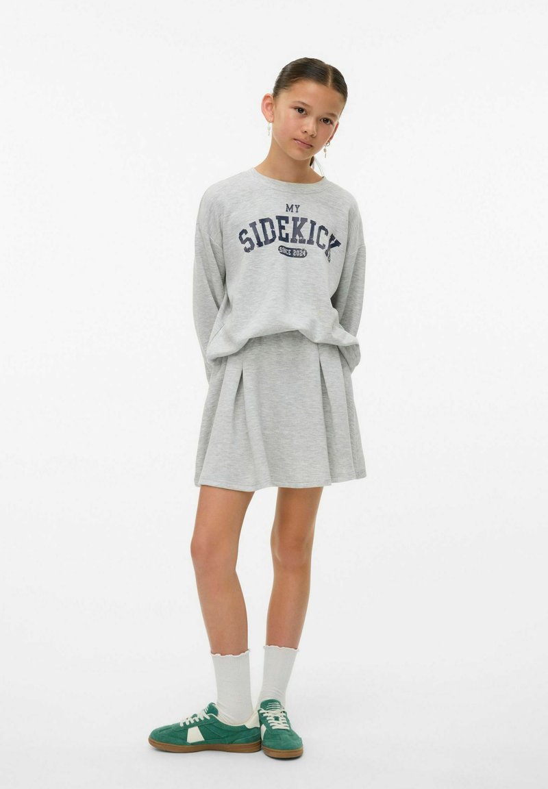 Sweatshirt gris avec le graphisme "MON PARTENAIRE", associé à une jupe plissée grise. Des chaussettes blanches et des baskets vertes complètent la tenue.