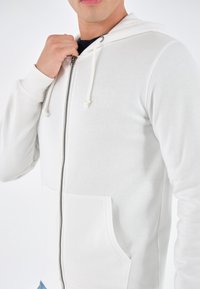 Homme portant un sweat à capuche blanc zippé avec cordons, main tenant la fermeture éclair près du cou, sur un fond uni.