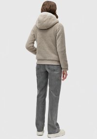 Hoodie en polaire beige avec une finition douce et texturée. Comprend une doublure intérieure blanche et une poche kangourou, associé à un jean droit gris.