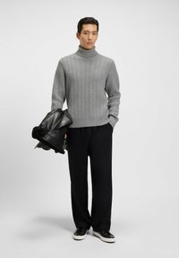 Grauer gestrickter Rollkragenpullover mit einem Zickzackmuster, kombiniert mit schwarzen weit geschnittenen Hosen und einer schwarzen Lederjacke, die über den Arm drapiert ist.