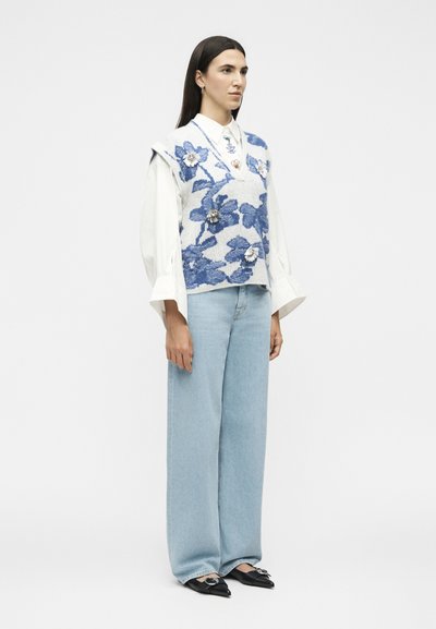 Femme portant une blouse blanche et un gilet en tricot bleu fleuri ample, un jean large bleu clair et des ballerines noires pointues avec une boucle argentée.