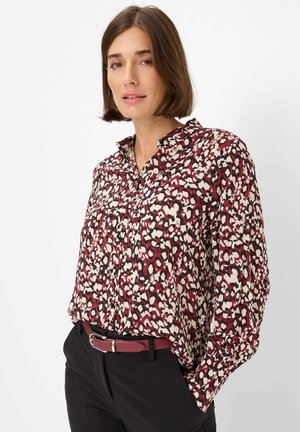 Blusa a maniche lunghe con stampa animalier maroon, crema e nera, con colletto e chiusura a bottoni; abbinata a pantaloni neri e una cintura coordinata.