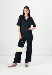 Completo a due pezzi in crepe blu navy con pieghe testurizzate. La donna tiene una borsa bianca con dettagli in pizzo. Muli grigi con tacco completano l'outfit.