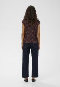 Žena kučeravých vlasov v hnedej blúzke s bodkami s krátkym rukávom, tmavomodrých džínsach s širokými nohavicami a čiernych topánkach, stojaca chrbtom.