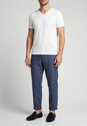 Muž, který má na sobě bílé tričko s krátkým rukávem a výstřihem do V, modré zakrácené kalhoty a černé nazouvací boty, stojí před jednobarevným pozadím.