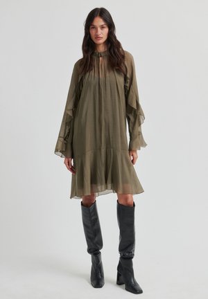 Olijfgroene, gelaagde jurk met gerimpelde mouwen en een hoge hals, gemaakt van lichtgewicht stof; gecombineerd met zwarte kniehoge laarzen.